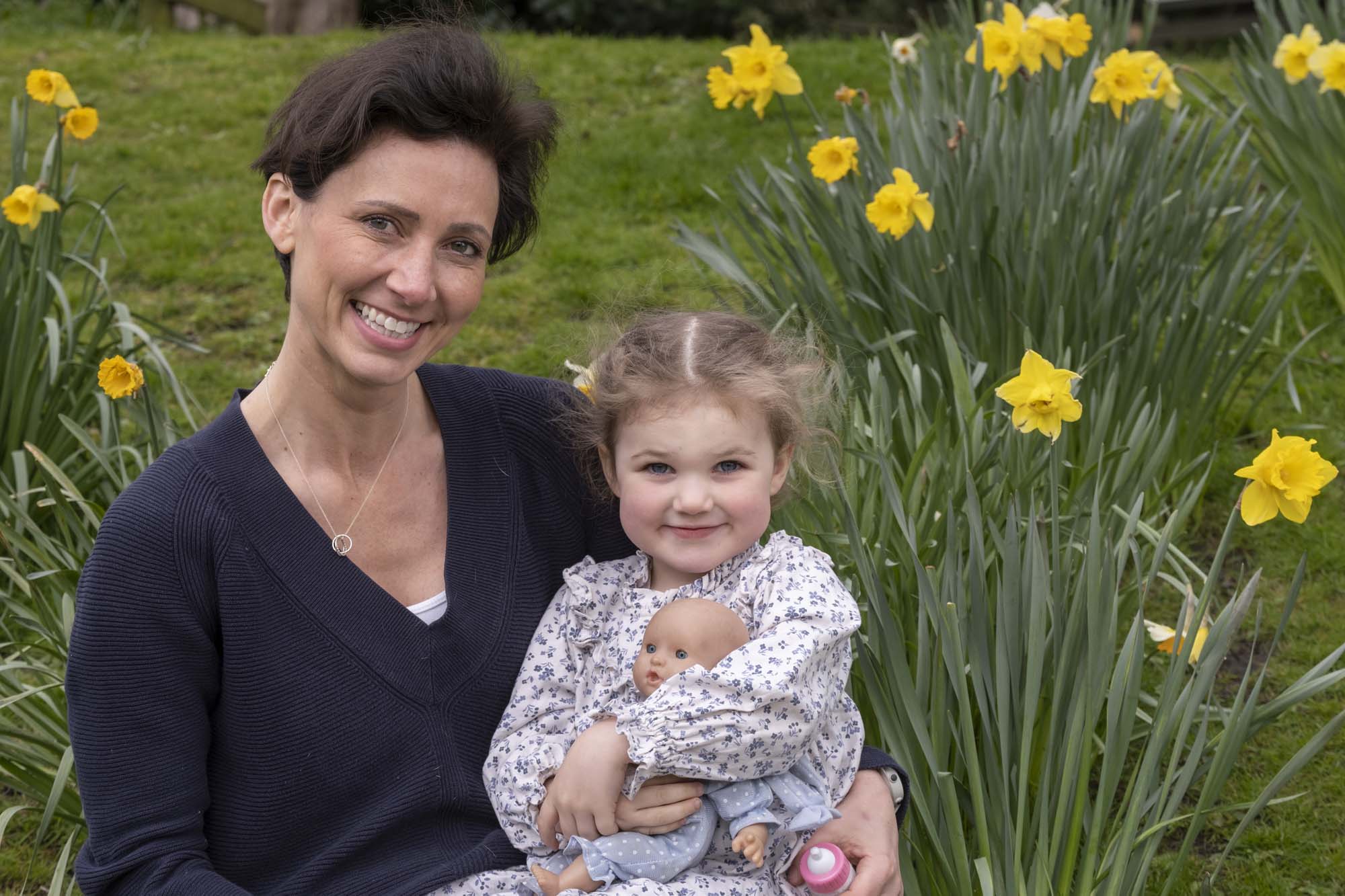 Victoria Robb and Matilda smile while sitting in a field surrounded by yellow flowers Matilda is holding a doll